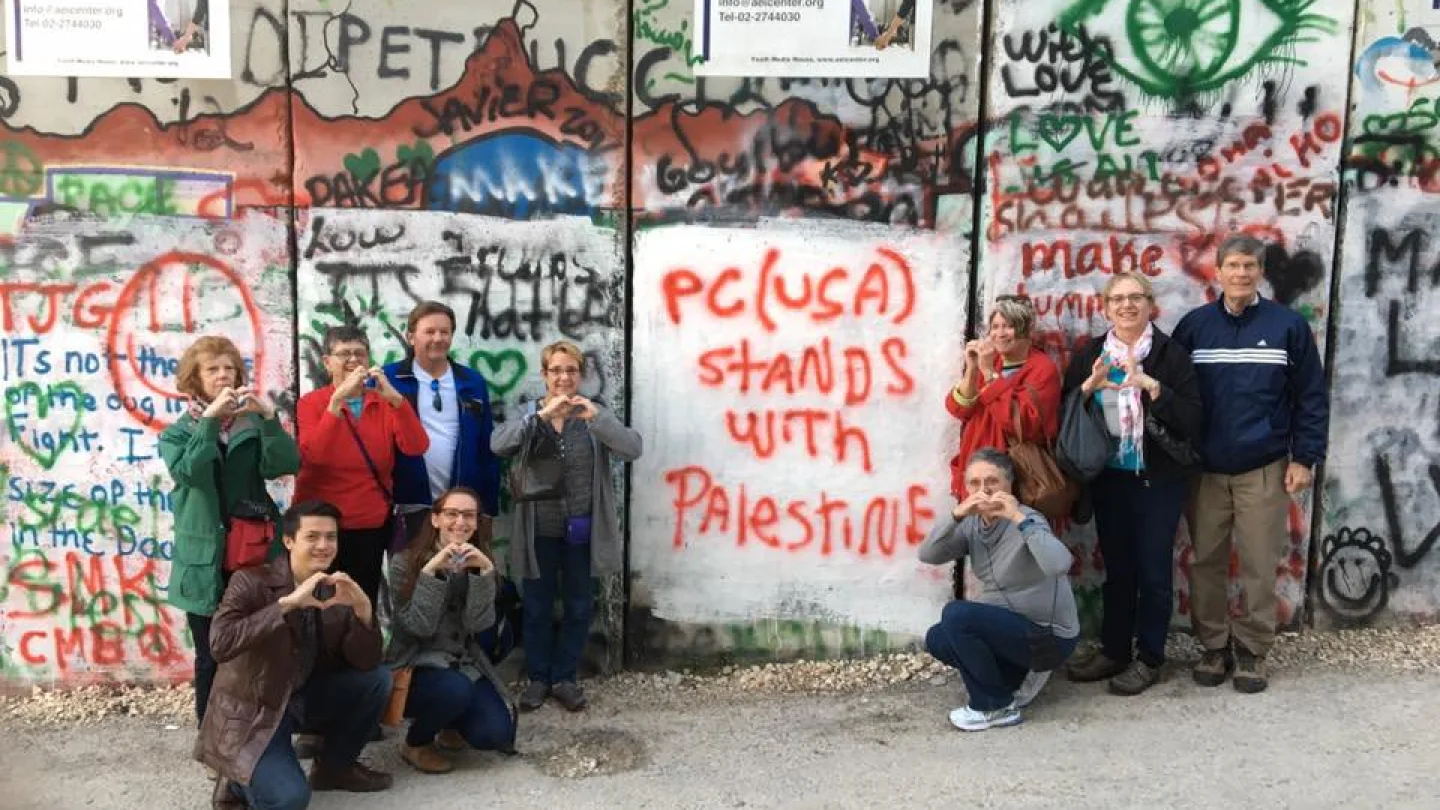 Members of the Presbyterian Church USA's Israel Palestine Mission Network pose in front of Israel's security barrier during one of their trips to the Holy Land. The graffiti on the barrier readers “PC(USA) stands with Palestine.” Source: Twitter.