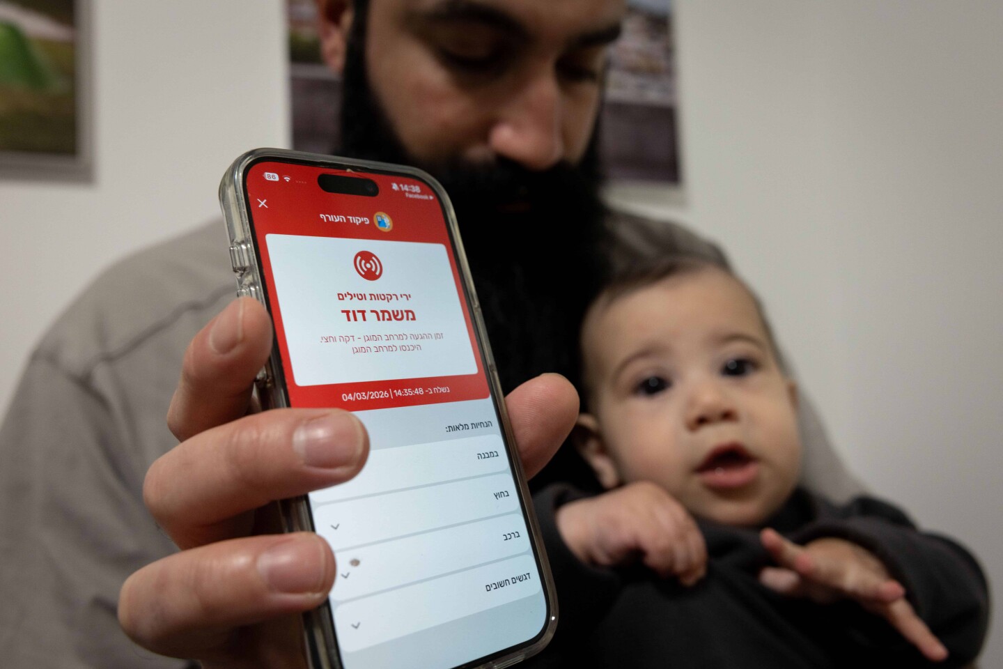 A man holding a baby shows a missile alert notification on her mobile phone as residents take cover indoors following missile fire from Iran toward Israel, in Mishmar David, March 4, 2026. Photo by Nati Shohat/Flash90.