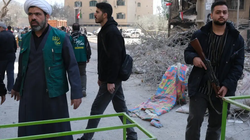 A Basij militiaman armed with an AK-47-style assault rifle stands guard outside a residential building in Tehran’s Shahid Borujerdi complex that was destroyed in a U.S.-Israeli missile strike during Operation Epic Fury, March 5, 2026. Photo by Kaveh Kazemi/Getty Images.