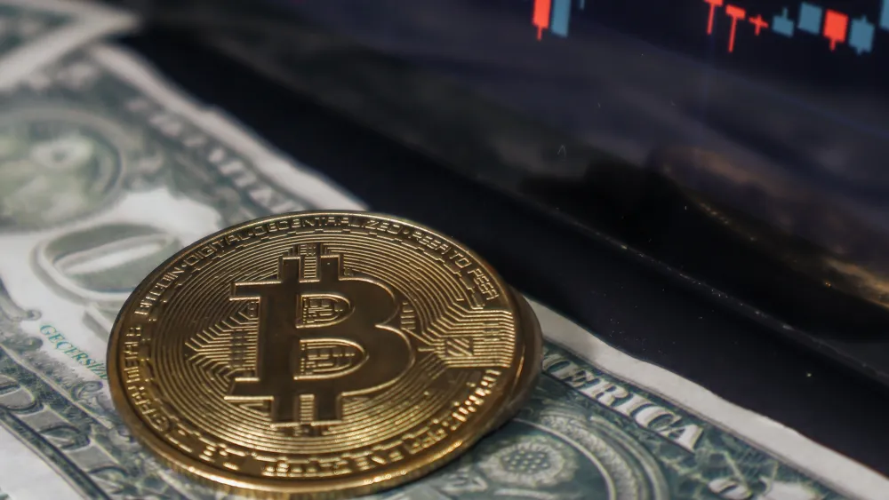 A cryptocurrency coin resting on a dollar bill with a market chart on a screen behind it. Credit: beyzahzah/Pexels.