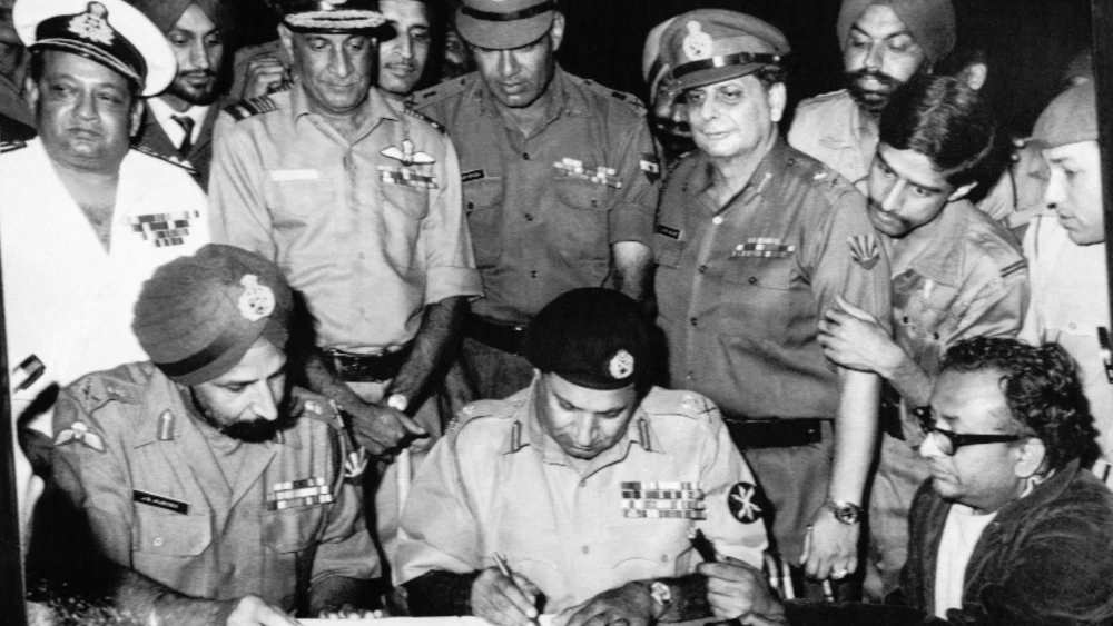 Lt. Gen. Amir Abdullah Khan Niazi signing the Instrument of Surrender under the gaze of Lt. Gen. Jagjit Singh Aurora. Standing immediately behind (from left) are Vice Adm. Nilakanta Krishnan, Air Marshall Hari Chand Dewan, Lt. Gen. Sagat Singh, Maj. Gen. Jack Farj Rafael Jacob (with Fg Offr SS Krishnamurthy peering over his shoulder). To their right, veteran newscaster Surojit Sen of All India Radio holds a microphone, Dec. 16, 1971. Credit: Indian Navy via Wikimedia Commons.