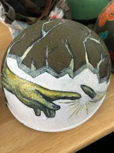A decommissioned IDF helmet painted by Asif Avrahami, an Egoz Unit reserve commander who took up painting after being wounded in combat. The artwork was inspired by a friend’s helmet, which was grazed by a sniper’s bullet and depicts God’s hand stopping the round. Credit: Courtesy.