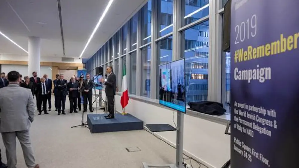 Italian Ambassador to NATO Claudio Bisogniero speaks at the NATO headquarters alongside a screen projecting #WeRemember photo via live stream from Auschwitz-Birkenau. Credit: NATO.