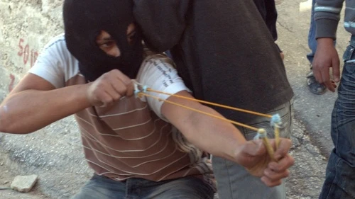 Click photo to download. Caption: In Silwan, an older balaclava-clad child with a slingshot fires off a stone at the cops, as cameras in front of him captured his “heroic” pose. Credit: B. Davidson.