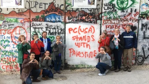Members of the Presbyterian Church USA's Israel Palestine Mission Network pose in front of Israel's security barrier during one of their trips to the Holy Land. The graffiti on the barrier readers “PC(USA) stands with Palestine.” Source: Twitter.