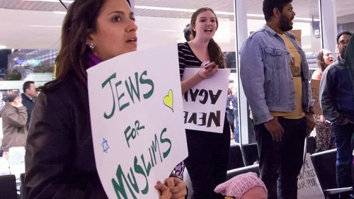At left, a protester at San Francisco International Airport holds a sign reading “Jews For Muslims” during a demonstration against the Trump administration’s temporary travel ban affecting Muslim-majority countries Jan. 29, 2017. Credit: Kenneth Lu via Wikimedia Commons.