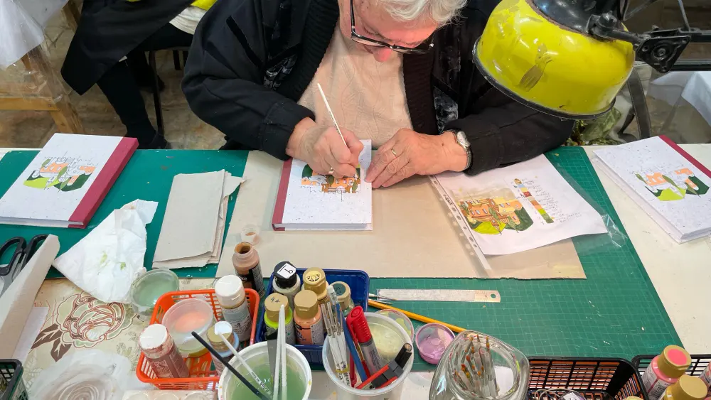 Olga, 77, hand illustrates a book cover at Yad LaKashish’s Paper Mache Workshop. Photo by Judith Segaloff.