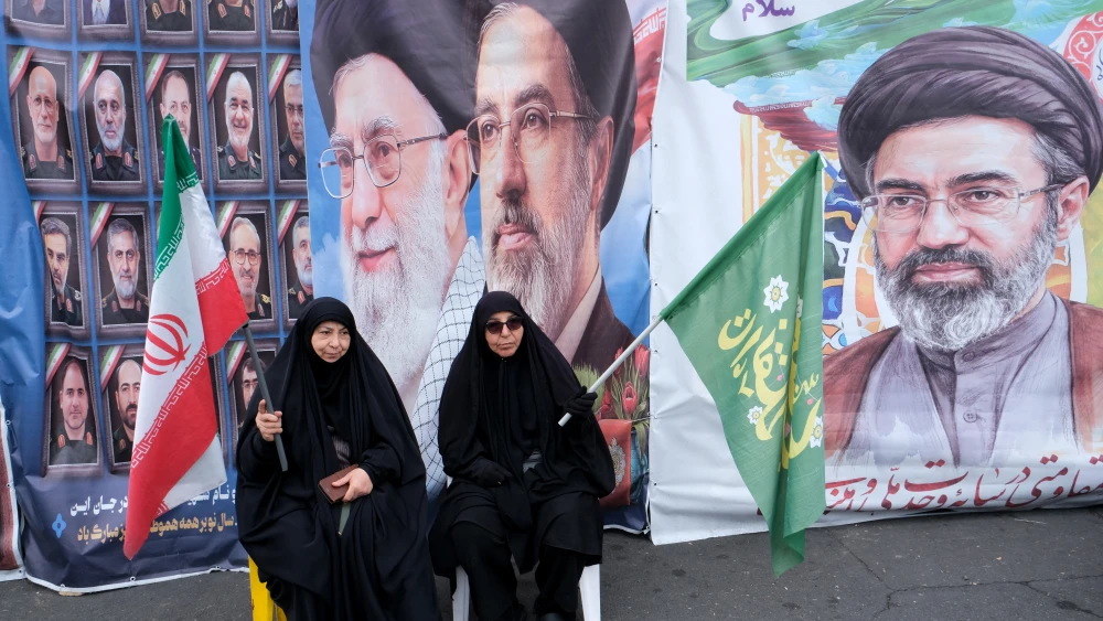 Two Iranian regime supporters sit on a bench in front of images of Ayatollah Ali Khamenei and his successor, Supreme Leader Mojtaba Khamenei, on the corner of Tehran's Revolution Square, March 28, 2026. Photo by Kaveh Kazemi/Getty Images.