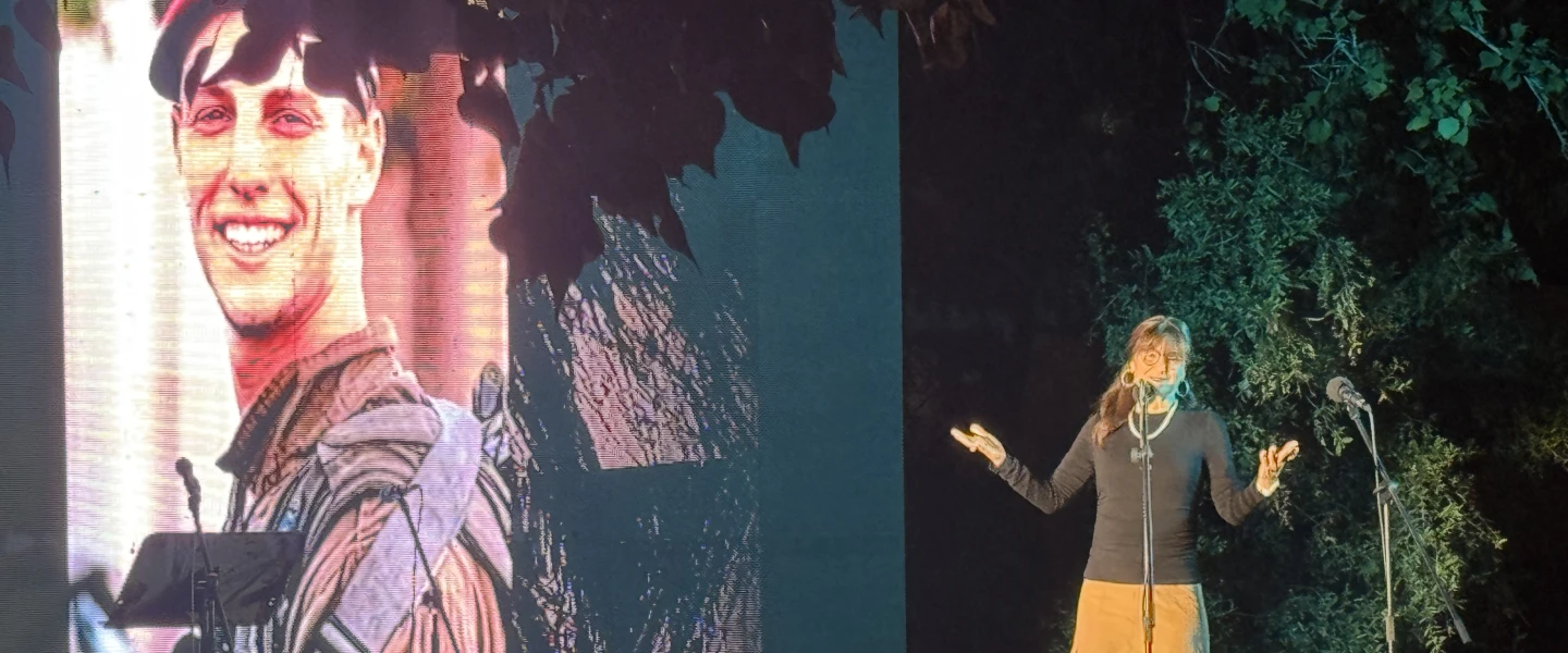 Jen Airley speaks at Kibbutz Nir Am’s Oct. 7 commemoration as a photograph of her late son, IDF paratrooper Binyamin Meir Airley, is projected behind her, Oct. 7, 2026. Photo by Howard Blas.
