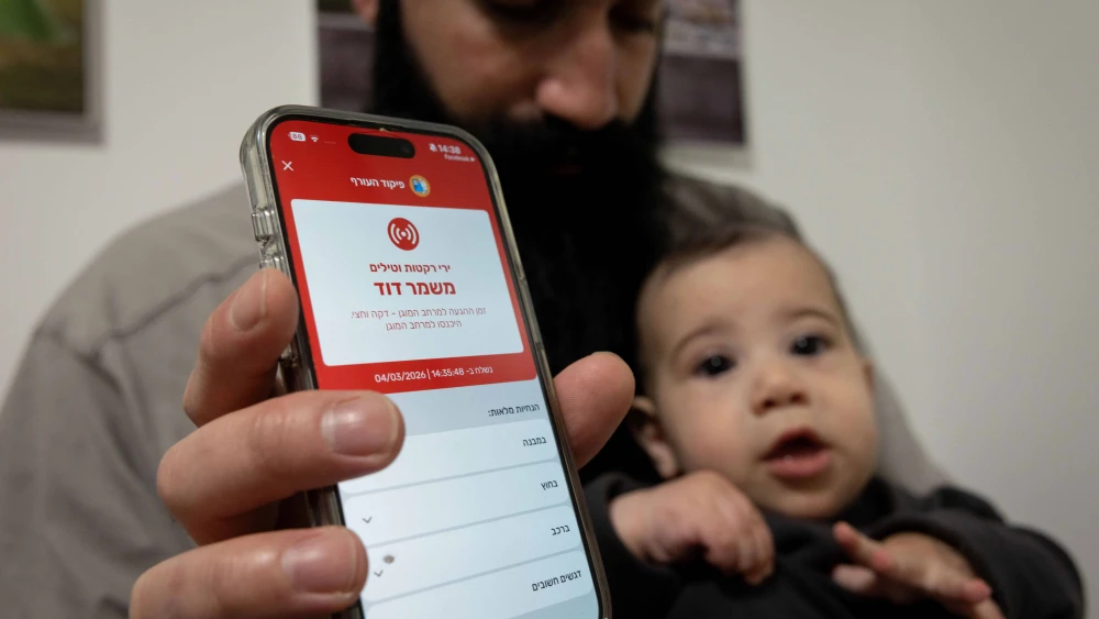 A man holding a baby shows a missile alert notification on her mobile phone as residents take cover indoors following missile fire from Iran toward Israel, in Mishmar David, March 4, 2026. Photo by Nati Shohat/Flash90.