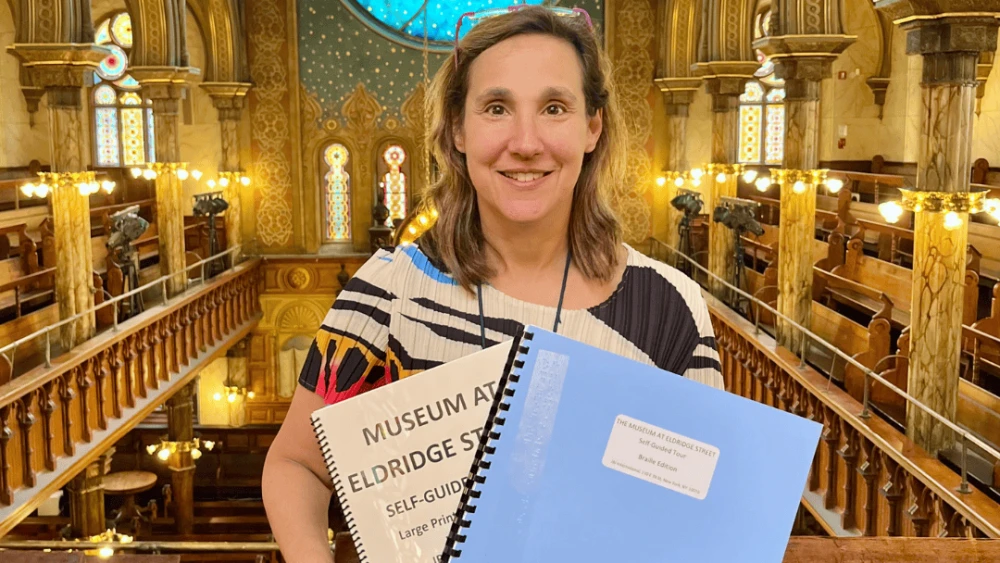 A woman holds a large print and braille self-guided tour booklet and stands inside a synagogue.